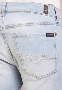 Ljust blå denimjeans med en lätt sliten bakficka, med en böjd sömfinish och en mörkblå tygetikett vid midjan.
