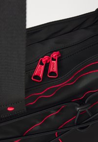 Black fabric bag featuring red zip pulls, with red stitching and a bungee cord detail. Smooth texture, functional design, and multiple compartments.