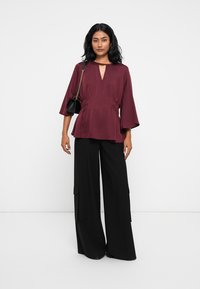 Blusa bordeaux con maniche a campana e scollatura a V, abbinata a pantaloni larghi neri con tasche laterali. Tessuto morbido e leggero.