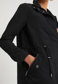 Zwarte lichtgewichtjas met een ritszak en een taillekoord. Heeft een kraag met drukknopen en een gladde stoftextuur.