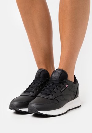 Mustat Reebok-kengät, joissa on sileä nahkavuori, valkoinen välikerros, tekstuuriset yksityiskohdat ja nauhakiinnitys. Varustettu pehmustetulla nilkkaremmillä ja kumipohjalla.