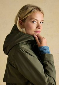 Oliwkowa, wodoodporna kurtka z kapturem, o gładkiej fakturze i denimowymi akcentami na mankietach, zaprojektowana w nowoczesnym kroju.