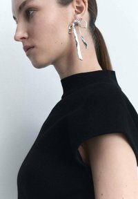 Orecchini astratti in argento con un design attorcigliato, pendenti sotto il lobo dell'orecchio. Top nero a collo alto con maniche corte e strutturate.