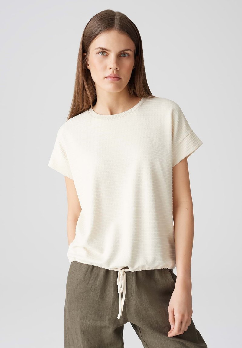 Helles beige T-Shirt mit kurzem Ärmel, strukturiertem Finish und elastischem Saum, kombiniert mit lockeren olivgrünen Hosen mit einem Kordelzug-Detail.
