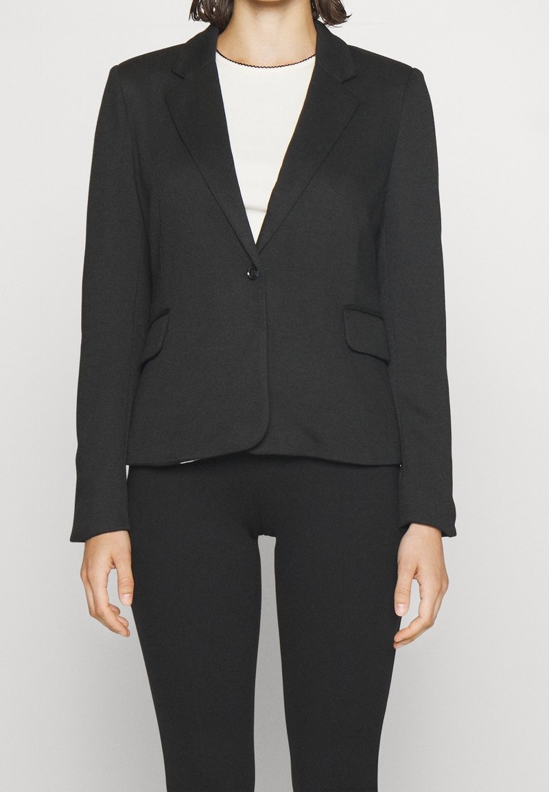 Personne portant un blazer noir ajusté avec un bouton sur un haut blanc, associé à un pantalon noir sur un fond clair uni.
