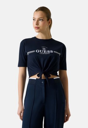 Mujer con top corto anudado Guess California en azul marino y pantalones de cintura alta con textura a juego, mirando de lado contra un fondo liso.