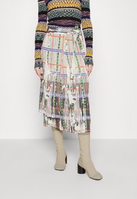Jupe midi plissée avec un motif vivant de lignes vertes, orange et bleues sur un fond blanc. Détail de taille nouée et design en couches.