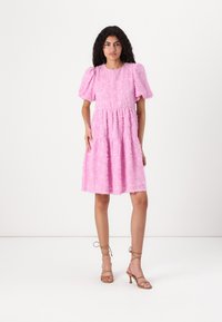 Vestido rosa, texturizado, com mangas bufantes, corpete ajustado e saia rodada. Combinado com sapatos de salto nude em um fundo neutro.