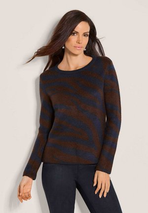 Vrouw met lang bruin haar die een bruin en donkerblauw gestreepte trui en een donkere broek draagt tegen een beige achtergrond.
