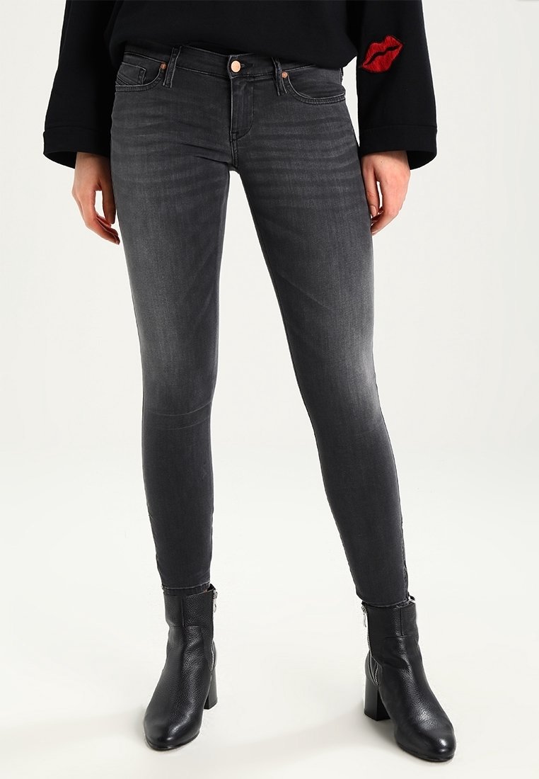 Person klädd i åtsittande urblekta svarta jeans, svarta ankelstövlar med blockklack och en svart långärmad topp med ett röd läppar patch på ärmen.