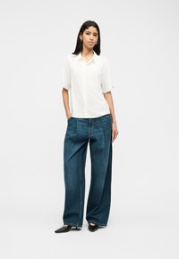 Γυναίκα που στέκεται φορώντας λευκό πουκάμισο με κοντό μανίκι και κουμπιά, μπλε τζιν με φαρδιά πόδια και μαύρα μυτερά παπούτσια, με τα χέρια στις τσέπες.