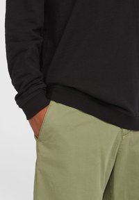 Maglietta nera a maniche lunghe con scollatura rotonda, abbinata a pantaloni verde oliva con tasche laterali e un tessuto liscio e leggero.