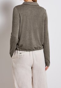 Haut à manches longues vert olive avec col, fabriqué en tissu doux. Il présente un ourlet inférieur froncé et est associé à un pantalon en lin blanc cassé.