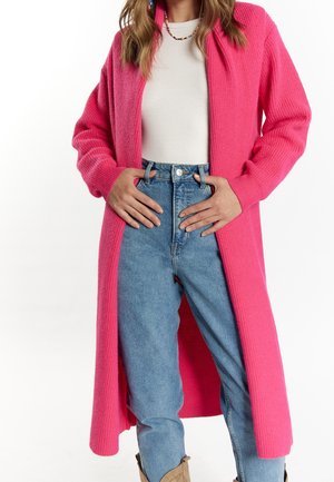 Femme portant un long cardigan rose vif par-dessus un top blanc, un jean bleu taille haute, un collier de perles colorées et des boucles d'oreilles.
