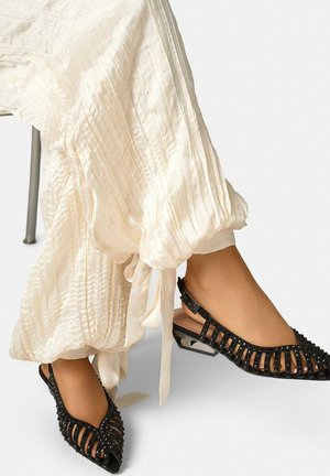 Pies con zapatos planos slingback negros con tachuelas y lados abiertos, combinados con pantalones holgados plisados de color crema atados en los tobillos.