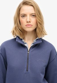 Granatowa bluzka z suwakem, wykonana z miękkiej bawełny, z wysokim kołnierzem, ściągaczami i haftem "SUPERDRY" na piersi.