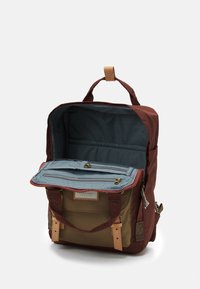 Brūns un olīvzaļš mugursomā ar atvērtu galveno nodalījumu, zilu rāvējslēdzēja organizatora atloku un ādas siksnām, attēlota uz balta fona.