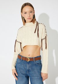 Maglione cropped a treccia crema con dettagli in frange marroni, abbinato a jeans blu a vita alta e una cintura marrone.