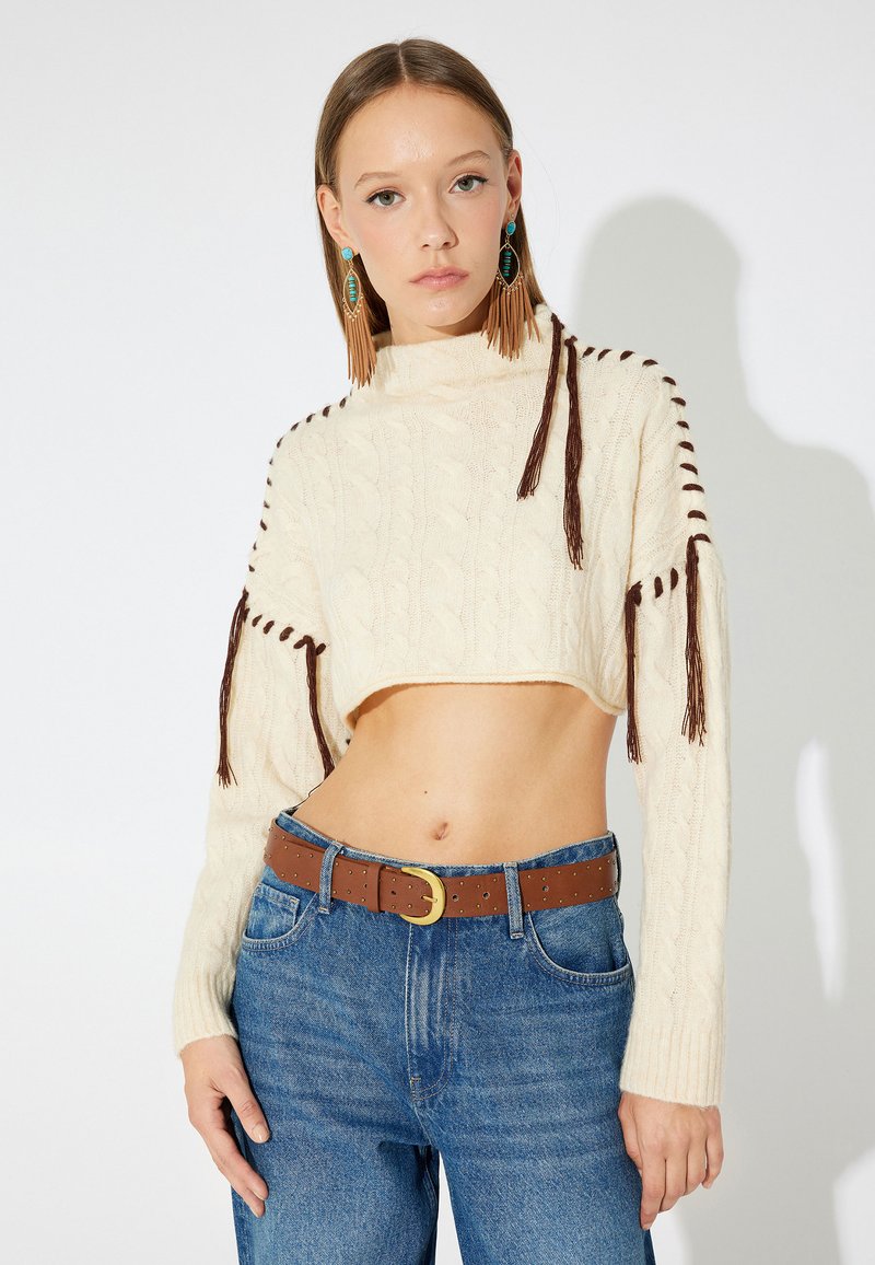 Maglione cropped a treccia crema con dettagli in frange marroni, abbinato a jeans blu a vita alta e una cintura marrone.