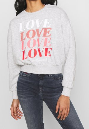 Femme portant un sweat-shirt court gris clair avec le mot « LOVE » répété en blanc, rose et rouge, et un jean bleu foncé ajusté.