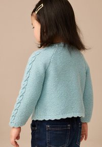 Cardigan lavorato a maglia di un azzurro chiaro con un motivo a cavi testurizzati sulle maniche e un orlo a scallop, abbinato a jeans scuri.