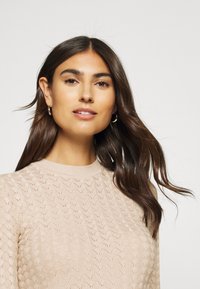Light beige knitted sweater featuring a textured wave pattern, ribbed crew neckline, and long sleeves. Model wears small hoop earrings.