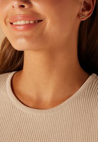 Gouden oorbellen in de vorm van kleine strikjes, gedragen met een gebreide beige top met een ronde halslijn. De huidtint is warm en glad.