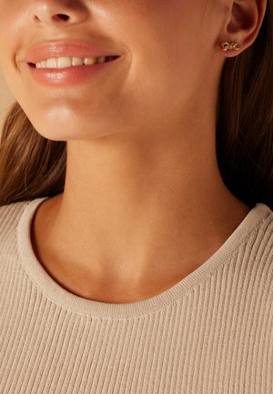 Gouden oorbellen in de vorm van kleine strikjes, gedragen met een gebreide beige top met een ronde halslijn. De huidtint is warm en glad.