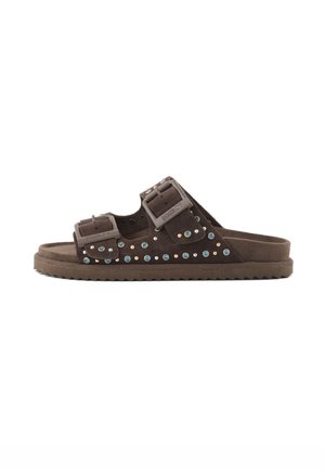 Sandalia plana de ante marrón con dos correas ajustables con hebillas adornadas con tachuelas azules y doradas, y una suela estriada.