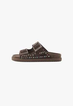 Sandalia plana de ante marrón con dos correas ajustables con hebillas adornadas con tachuelas azules y doradas, y una suela estriada.