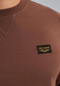 Sweatshirt marron avec un col rond, doté d'un tissu texturé. Inclus une étiquette rectangulaire noire avec du texte doré et un emblème.
