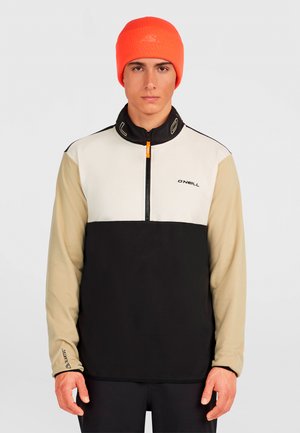 Man wearing a color-block quarter-zip pullover with beige sleeves, black body, white chest, and an orange beanie hat.