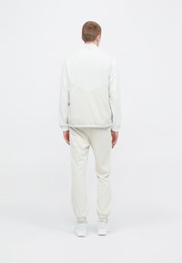 Chándal de color claro que incluye una chaqueta con cremallera y diseño en chevron con puños elásticos, combinada con pantalones entallados y zapatillas.