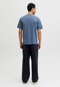 Man som står vänd bortåt iklädd en blå t-shirt med kort ärm, lösa svarta byxor och ljusa skor mot en enfärgad bakgrund.