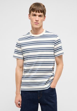 Junger Mann, der ein weißes T-Shirt mit marineblauen horizontalen Streifen und dunkelblaue Jeans trägt, steht mit einer Hand in der Tasche.