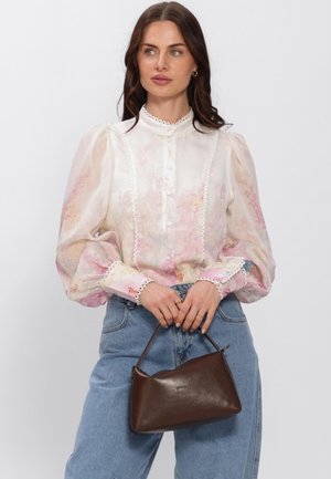 Vrouw die een doorschijnende bloemenblouse met pofmouwen en blauwe spijkerbroek draagt, en een kleine bruine leren handtas vasthoudt voor een witte achtergrond.