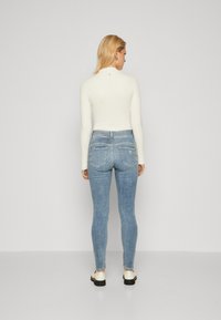Hvid langærmet top med høj hals, båret med blå skinny jeans med baglommer og et lille logodetail.