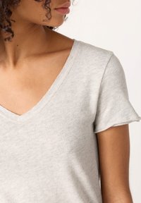 Sieva ar cirtainiem matiem, valkājot gaiši pelēku V veida kakla īsroku krekla, redzama no kakla līdz augšdelmam, skatās pa kreisi uz balta fona.