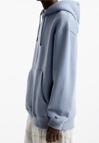 Persona vistiendo una sudadera oversized de color azul claro con bolsillo delantero y cordones, combinada con pantalones beige cosidos, contra un fondo blanco.