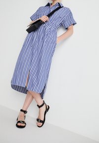 Blauw-wit gestreepte overhemdjurk met korte mouwen, een kraag, zijzakken en een voorste split. Gecombineerd met zwarte sandalen met gespsluiting.