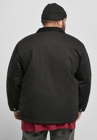 Giacca nera isolata con colletto, aperture posteriori e bottoni a pressione. Indossata sopra una camicia rossa, presenta una vestibilità comoda e un tessuto resistente.