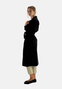 Czarny płaszcz trench z długimi rękawami, z paskiem w talii i kołnierzykiem koszulowym, zestawiony z jasnobeżowymi spodniami w kroju cropped i ciemnymi płaskimi butami.