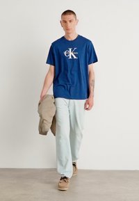 Camiseta azul marino con el logo blanco "CK", pantalones verde menta claros, chaqueta beige sobre el brazo y zapatos marrones. Modelo camina sobre un suelo neutro.