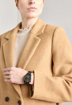 Czarny zegarek z siatkowym paskiem, z okrągłą tarczą i minimalistycznym designem, noszony na ręce spoczywającej na brązowym płaszczu w wzór rybiej ości.