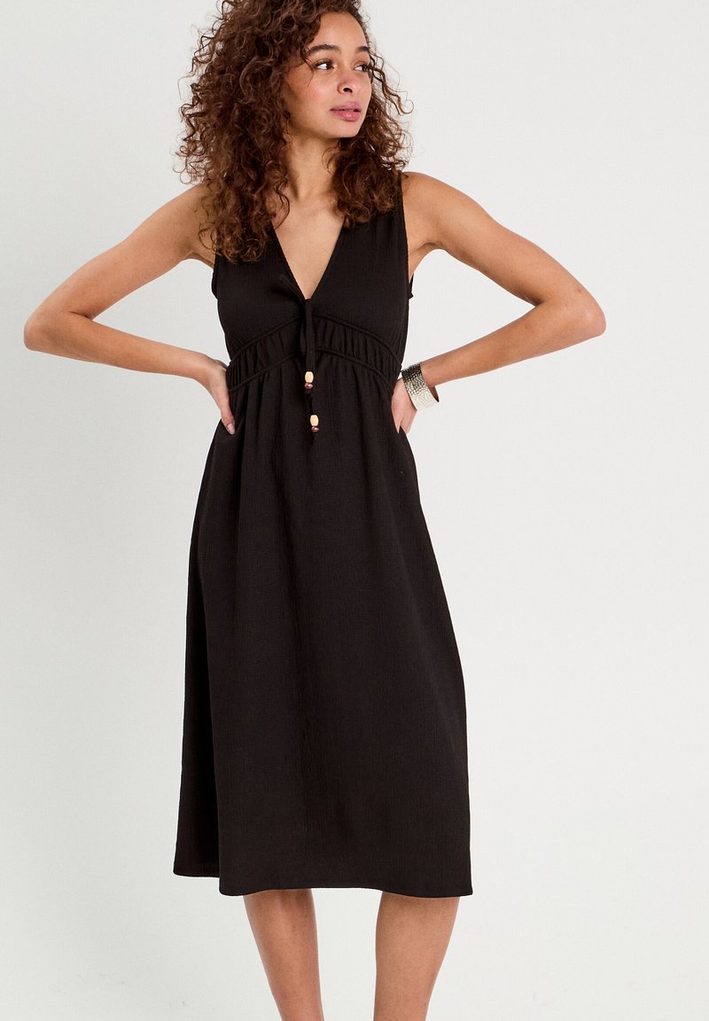 Robe noire sans manches avec un décolleté en V, taille froncée et perles décoratives. Fabriquée dans un tissu léger avec une finition texturée.