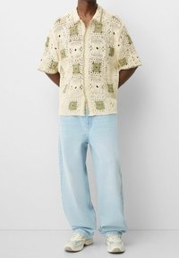 Camiseta de crochet color crema con patrones cuadrados verdes, mangas cortas y cuello, combinada con vaqueros de corte holgado azul claro y zapatillas deportivas.