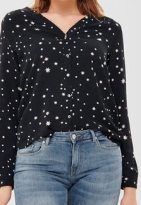 Svart skjorta med stjärnmönster i vitt, med V-ringad hals och knäppning fram. Långa ärmar och en lös passform. Bärs med ljusblå jeans.