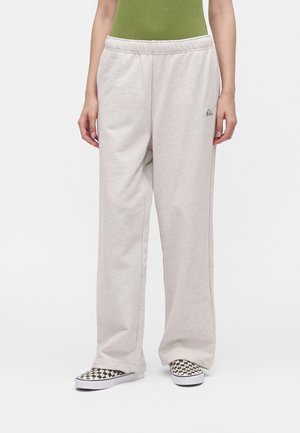 Personne portant un pantalon de survêtement ample beige clair et des chaussures slip-on à carreaux noir et blanc, debout sur un fond blanc.