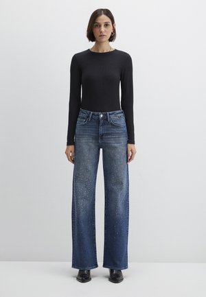 Weite Denim-Jeans mit einer verblassten blauen Waschung, verziert mit verstreuten Strassstein-Applikationen. Kombiniert mit einem schwarzen Langarmoberteil.