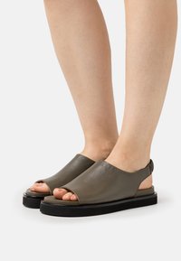 Fötter i öppna sandaler med bakrem i mörk olivgrön läder och tjocka svarta sulor på vit bakgrund.